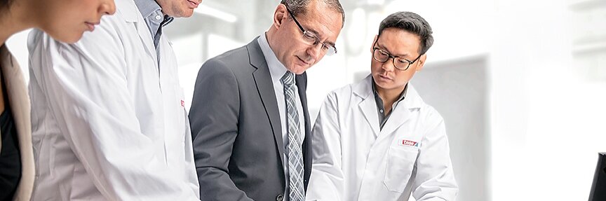 Four people are gathered around a table in a bright office. Two are wearing lab coats, one is in a suit, and one is partially visible. They are focused on documents laid out on the table, which include diagrams labeled with tesa tape. A laptop displaying pertinent data is open nearby.