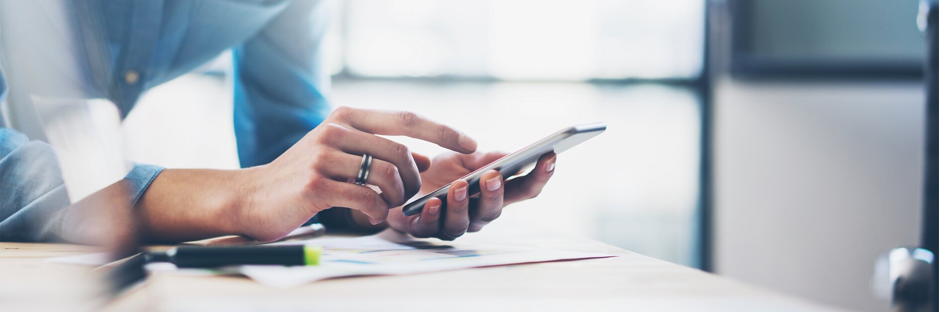 A person wearing a blue shirt is standing at a desk, using a smartphone. Their fingers are tapping the screen. Papers and a pen are visible on the desk, along with some tesa tape, with a laptop partially in the foreground. The setting is well-lit. (This text has been generated by AI)