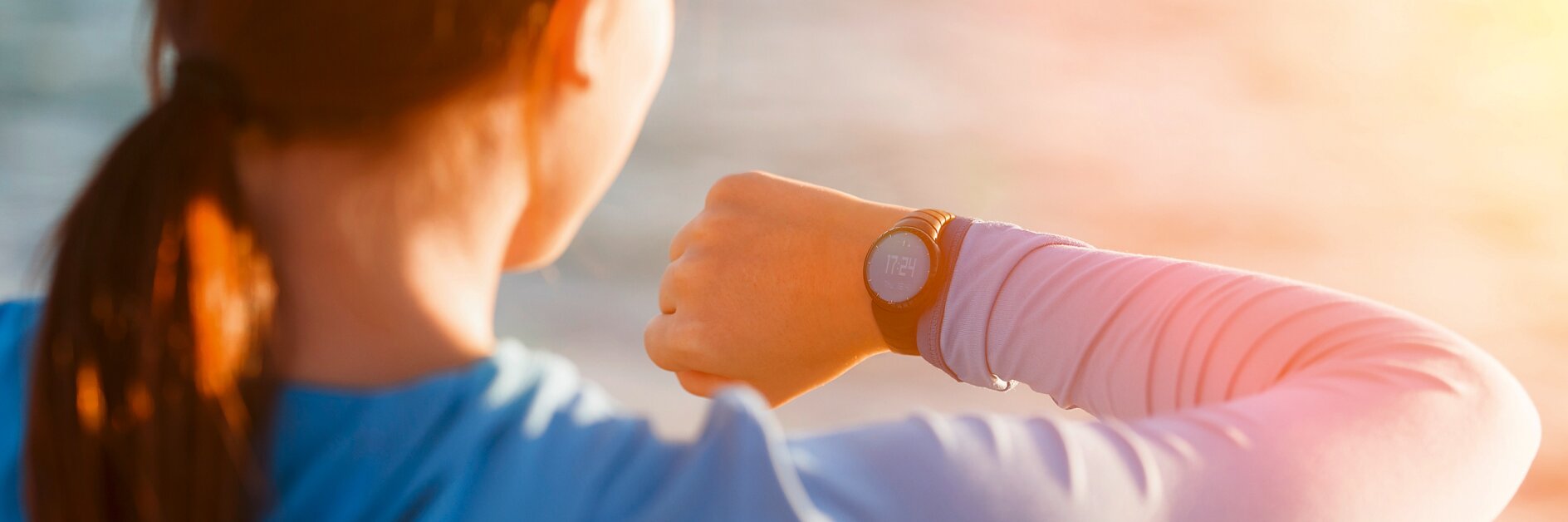 A person with reddish-brown hair, wearing a blue long-sleeve shirt, is looking at a smartwatch on their wrist. The background is blurred with sunlight and appears to be outdoors near water. (This text has been generated by AI)
