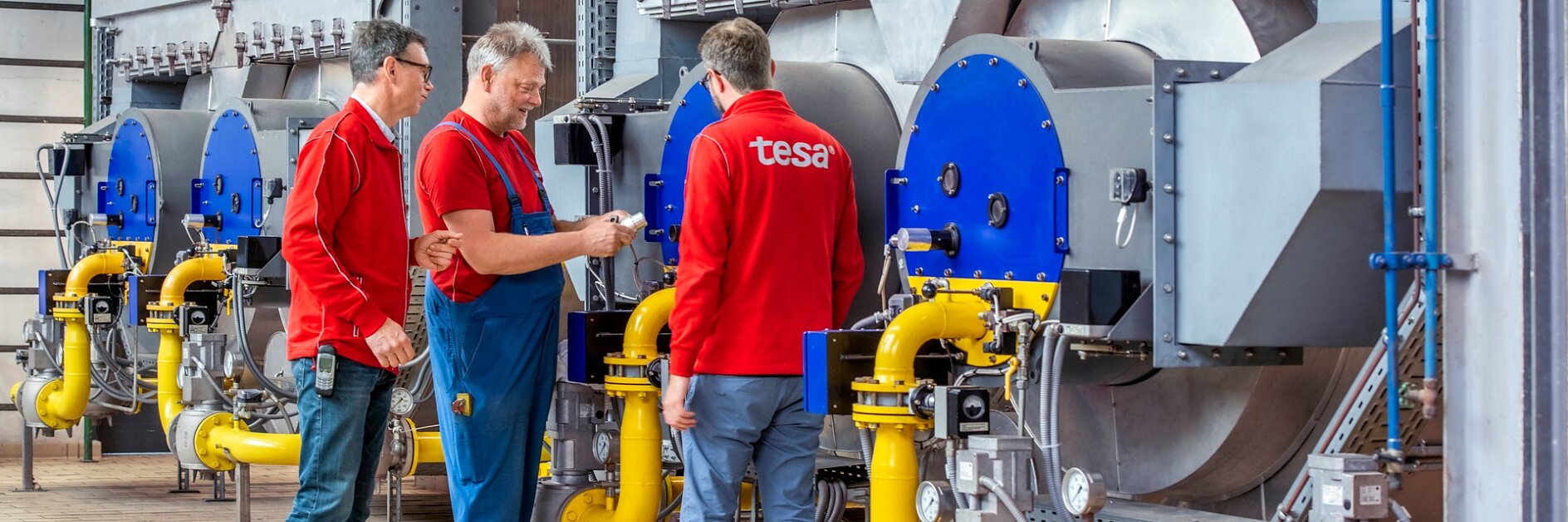 Three men stand in front of large industrial machinery with blue and silver components. They are wearing red shirts and are having a discussion. Yellow pipes are connected to the machines, and the floor is paved with tiles. (This text has been generated by AI)