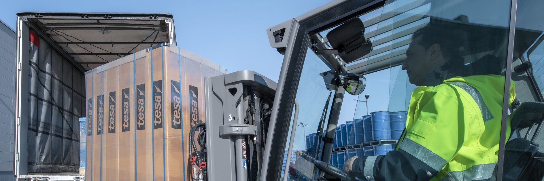 A forklift operator wearing a fluorescent yellow jacket loads a pallet of wrapped goods labeled Fessi into the back of a partially open truck. Blue barrels are visible in the truck. The scene takes place outdoors on a clear day, and everything is securely fastened with tesa tape. (This text has been generated by AI)