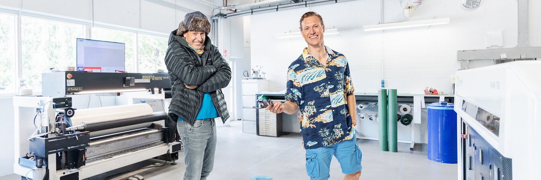 Two people stand in a bright workspace. One wears a beanie and puffy jacket, crossing their arms, while the other sports a Hawaiian shirt and shorts. A large printer is in the background along with some rolled materials. One of them looks serious, while the other is smiling. (This text has been generated by AI)