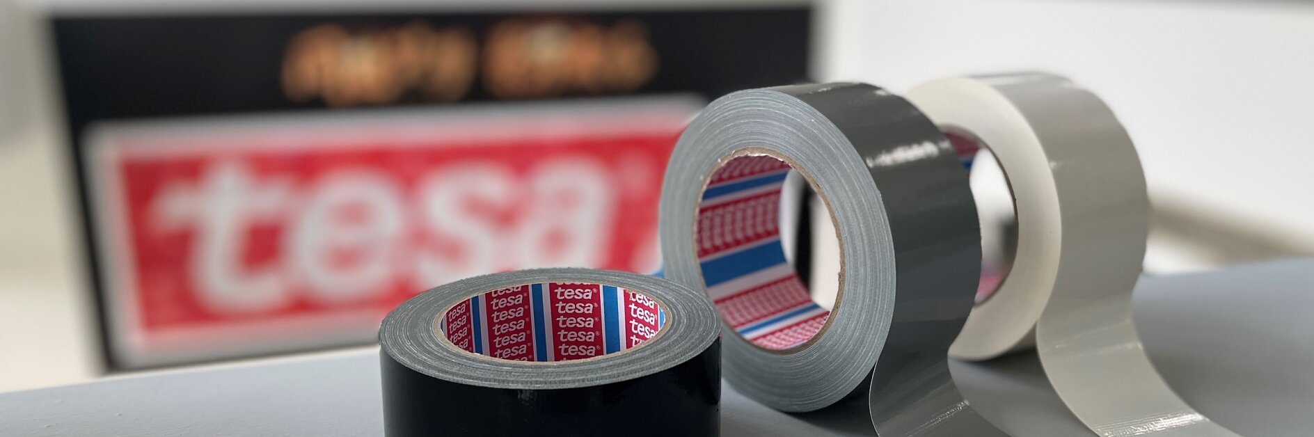 Three rolls of adhesive tesa tape are displayed: one black, one gray, and one white. The tesa tapes are positioned on a smooth surface, with a blurred tesa logo in the background. (This text has been generated by AI)