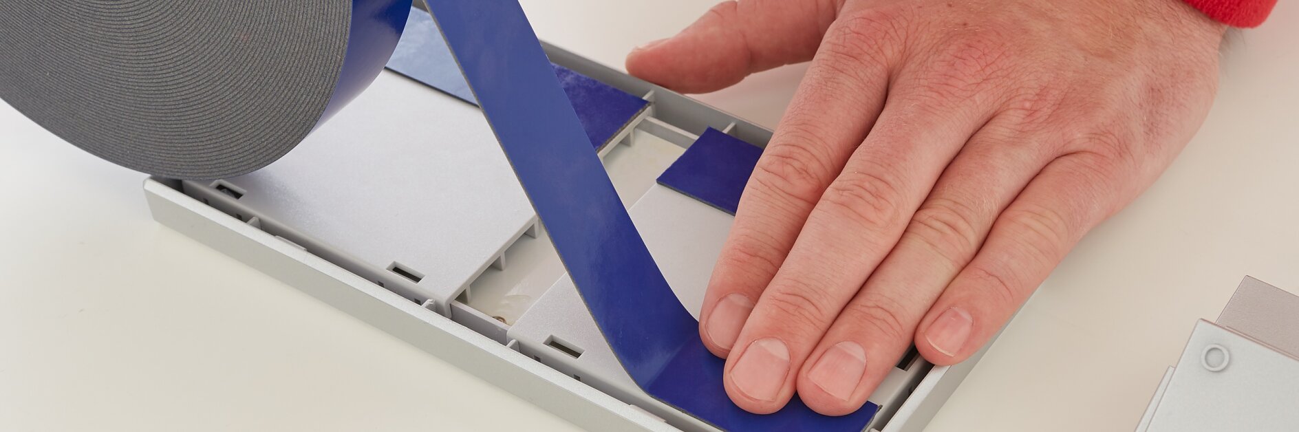 A person is applying blue tesa tape to a rectangular surface on a white table. The tesa tape is unrolled from a large roll, and the person uses their right hand to smooth it down. A red sleeve is visible on the persons arm. (This text has been generated by AI)