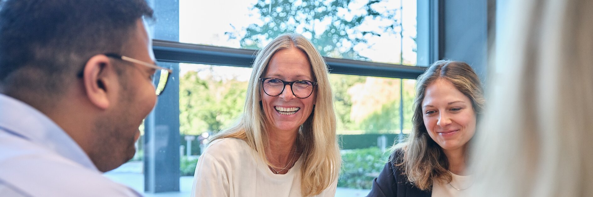 A group of four people sitting at a table in an office setting, engaging in conversation and enjoying a bowl of snacks. One person, wearing glasses, is smiling. A large window in the background shows a green outdoor view. (This text has been generated by AI)