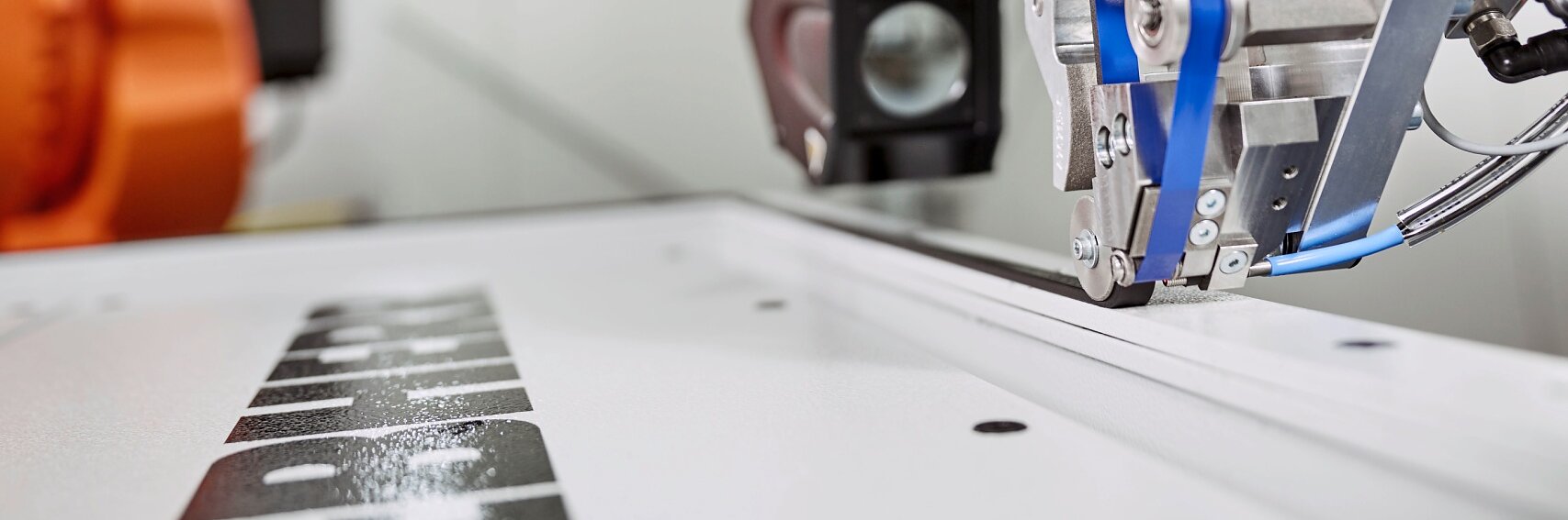 Close-up of industrial machinery in a manufacturing setting. The machine features metal components and blue tubing. Theres a spool of blue tesa tape material in the background, and a label on the machine reads “Mikan Technic.”. (This text has been generated by AI)