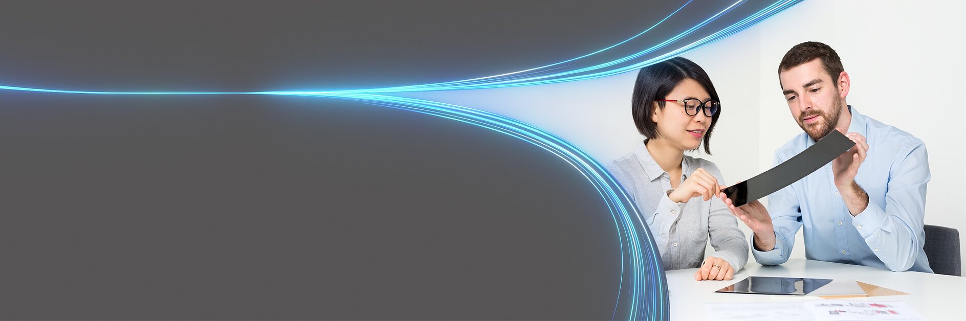 Two people are examining a flexible screen or device, sitting at a table with a tablet, papers, and tesa tape. The background features a gradient with blue neon lines curving across a grey space on the left, creating a futuristic look. (This text has been generated by AI)