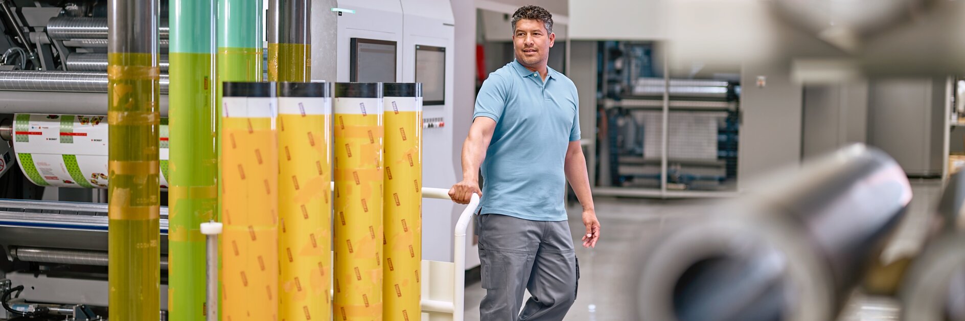 A man in a light blue polo shirt and gray pants stands in an industrial setting, surrounded by large rolls of colorful film. He rests one hand on a display of yellow and green rolls of tesa tape. Machinery and equipment are visible in the background. (This text has been generated by AI)