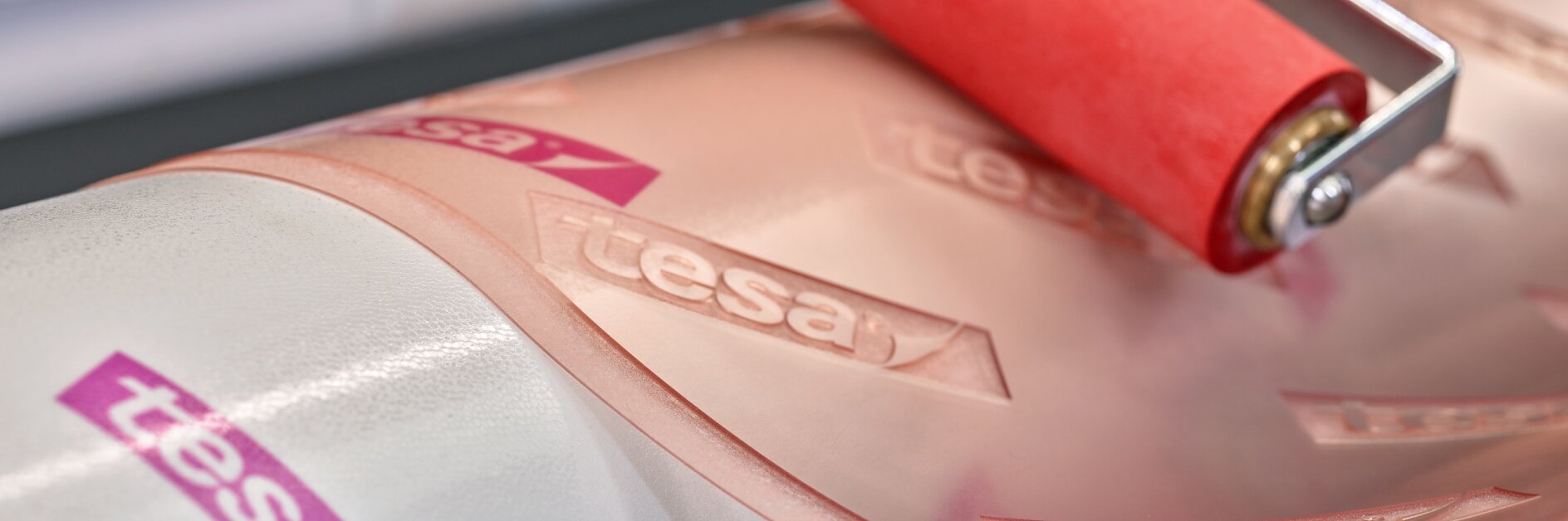Close-up of a hand using a red roller on a clear adhesive film marked with the tesa tape logo. The film is being applied to a white cylindrical surface.