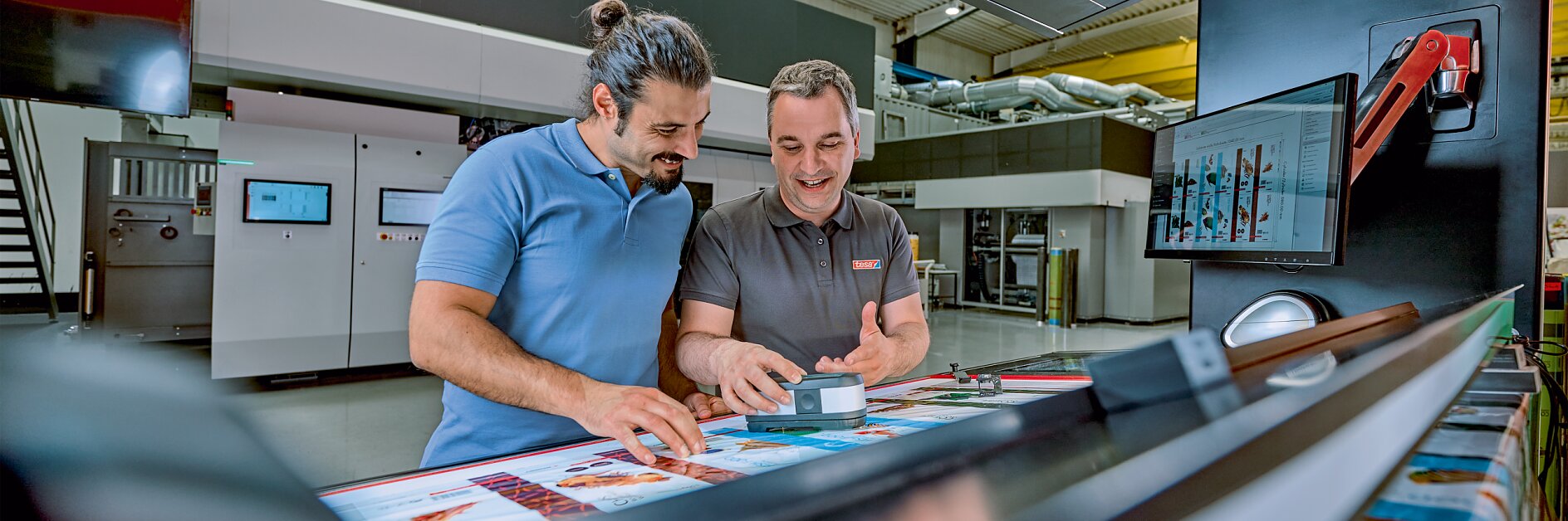 Two men are standing in a modern printing facility, examining colorful printed materials on a large machine. One man is holding a small box of tesa tape and smiling. There are screens and equipment in the background. (This text has been generated by AI)