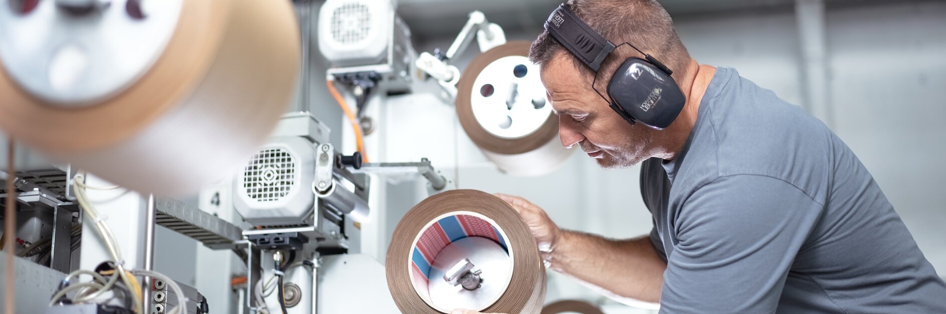 A man wearing ear protection inspects a roll of material in an industrial setting. Various machines with similar rolls of tesa tape are visible in the background. He is focused on examining the roll closely. (This text has been generated by AI)