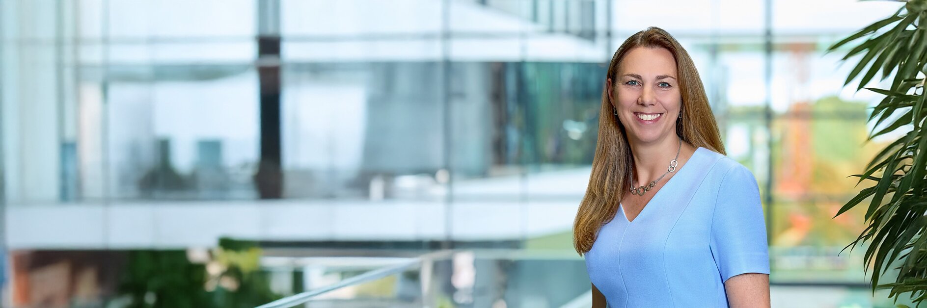 A woman with long brown hair, wearing a light blue short-sleeve top and a necklace, stands indoors by glass windows and green plants, smiling. (This text has been generated by AI)